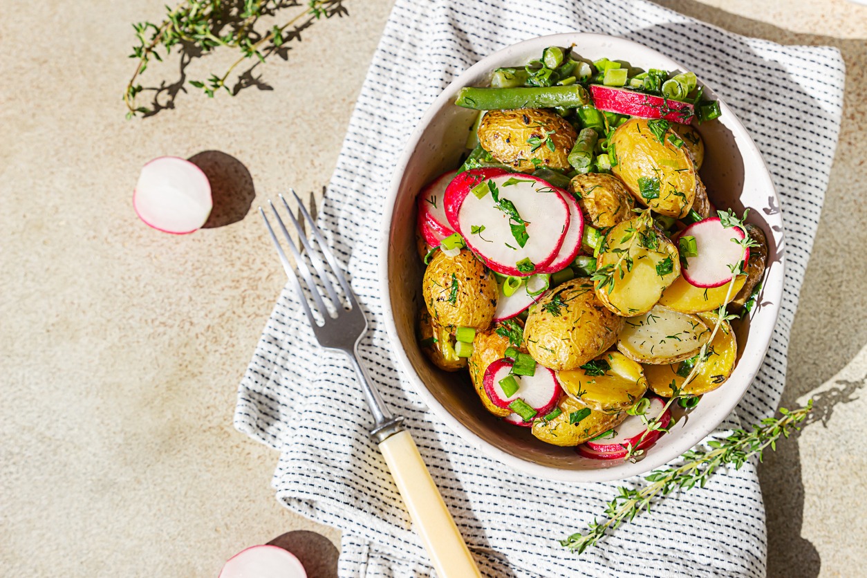 Tasty hot potato salad with green beans, fresh radishes and herbs dressing with olive oil and mustard sauce, light concrete background. Top view.