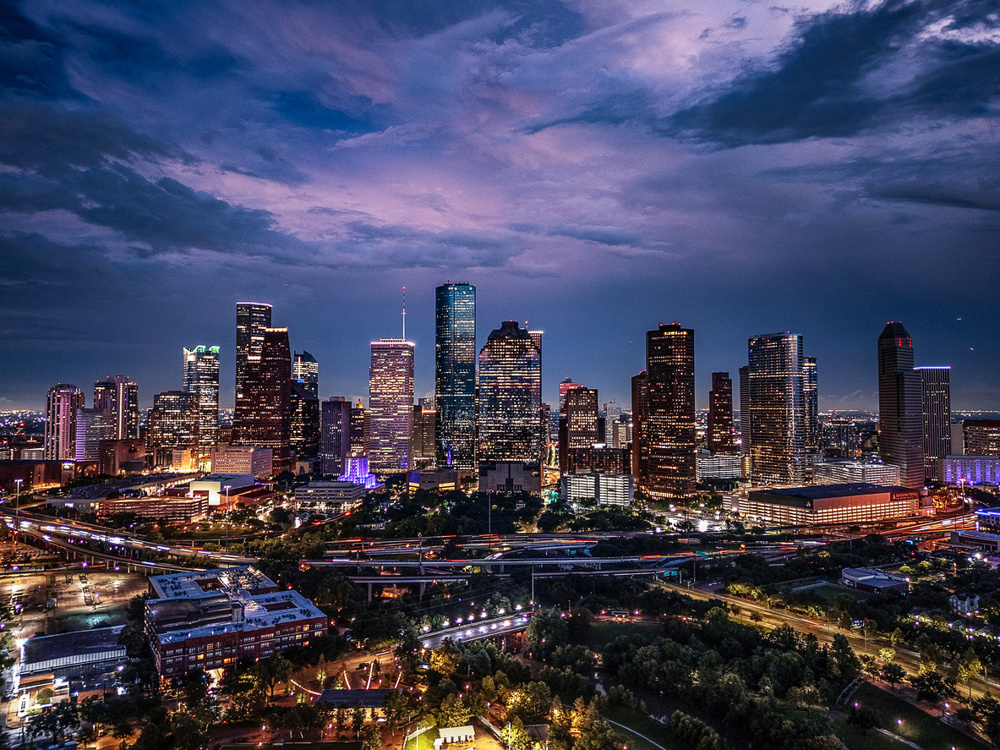 Drone aerial image of downtown Houston, Texas captured at twilight. The photograph highlights the illuminated skyscrapers, highways, and cityscape under a dramatic evening sky. A vibrant view of one of the largest cities in the United States and a major economic hub