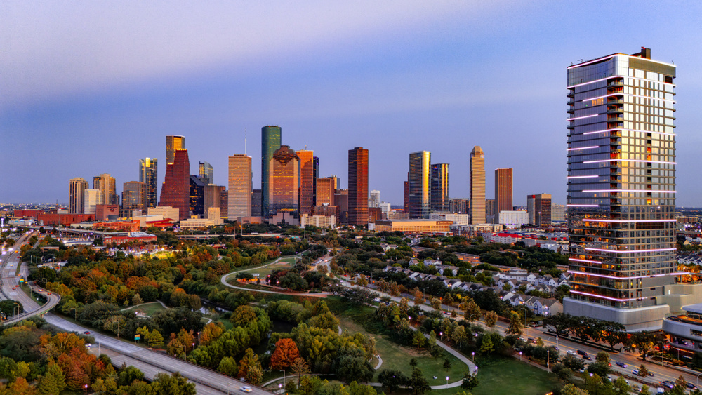 A high-resolution drone photograph of the Houston Texas skyline at sunset. The downtown towers glow with warm evening light while tree-lined parks and freeways stretch across the foreground. This elevated aerial panorama captures the city’s architecture, urban growth, and vibrant colors at golden hour