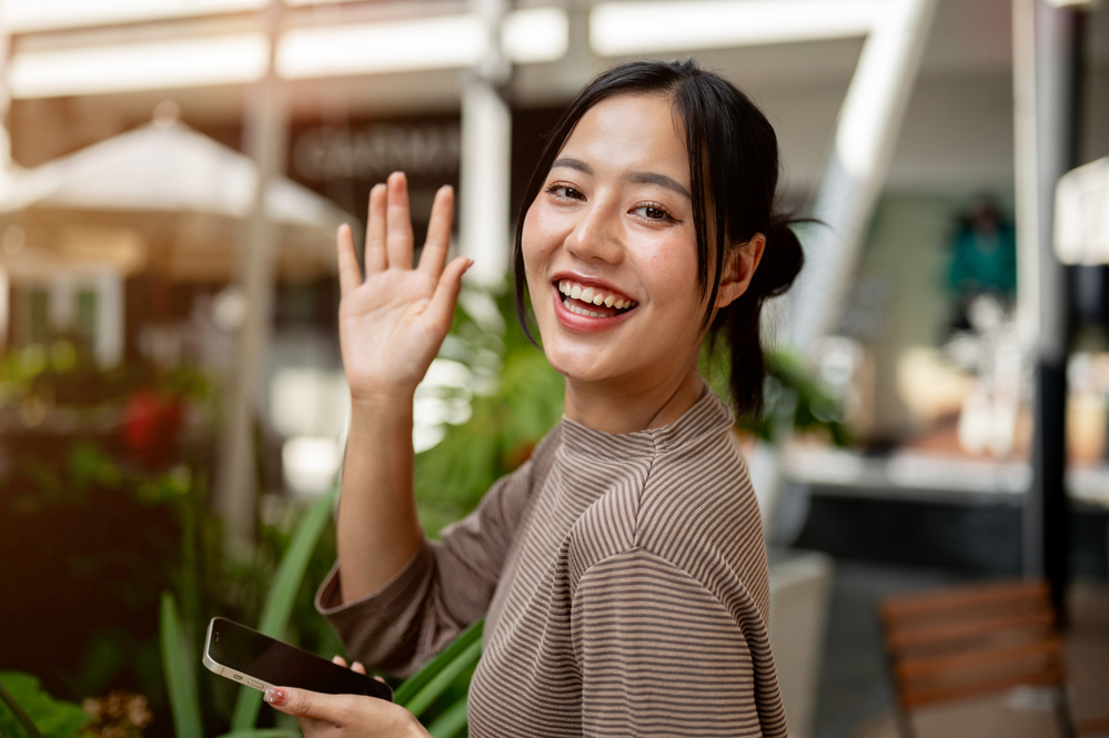 A portrait of a beautiful, positive Asian woman in casual clothes stands outdoors with a smartphone, waves and smiles at the camera. people, lifestyles, and wireless technology